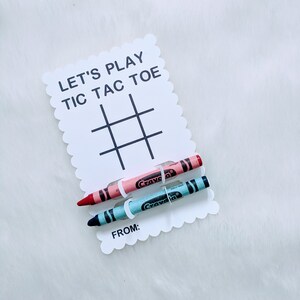 May include: A white card with the text "Let's Play Tic Tac Toe" and a tic-tac-toe grid. Two crayons, one pink and one blue, are attached to the card with white string.
