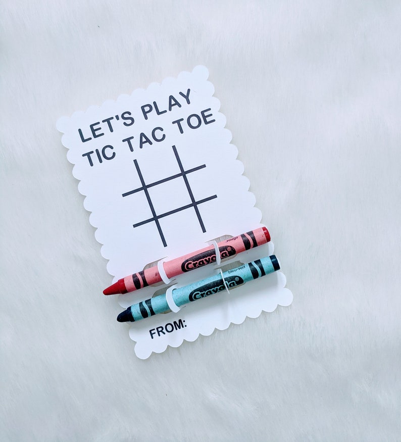 May include: A white card with the text "Let's Play Tic Tac Toe" and a tic-tac-toe grid. Two crayons, one pink and one blue, are attached to the card with white string.