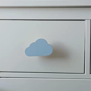 May include: A white drawer with a light blue cloud-shaped handle. The handle is in the centre of the drawer, which is part of a white dresser. The drawer is closed.