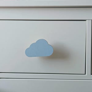 May include: A white drawer with a light blue cloud-shaped handle. The handle is in the centre of the drawer, which is part of a white chest of drawers. The cloud handle adds a playful element to the furniture.
