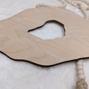 May include: A wooden cutout in the shape of an agate with a hole in the center. The cutout is hanging from a white beaded garland with a brown tassel.