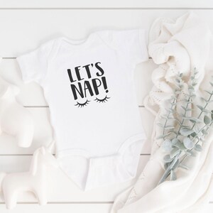 Puede incluir: Un body blanco para bebé con texto negro que dice "Let's Nap!" con dos pestañas negras encima del texto.