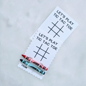 May include: Two white cards with scalloped edges and black text that reads "Let's Play Tic Tac Toe" and a tic-tac-toe grid. Two crayons, one pink and one blue, are attached to the cards with a clear plastic band.