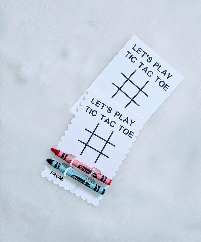 May include: Two white cards with scalloped edges and black text that reads "Let's Play Tic Tac Toe" and a tic-tac-toe grid. Two crayons, one pink and one blue, are attached to the cards with a clear plastic band.