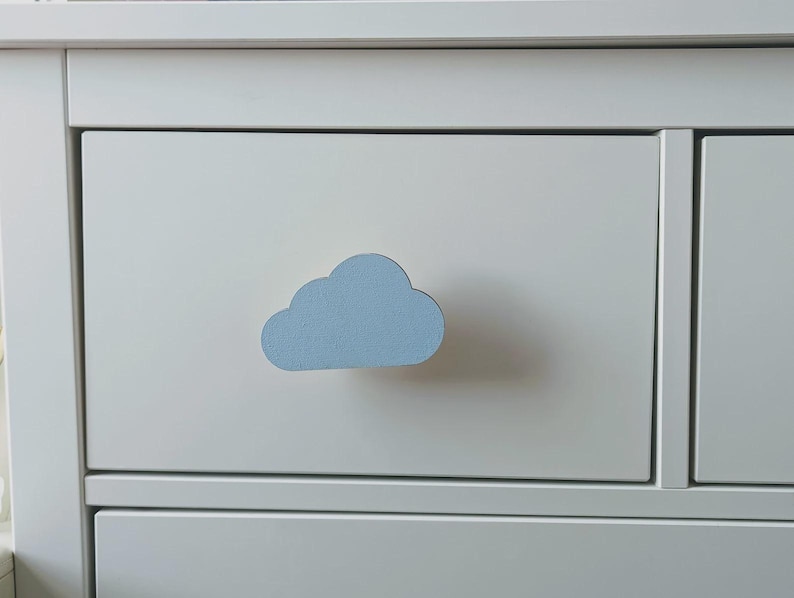 May include: A white drawer with a light blue cloud-shaped handle. The handle is in the centre of the drawer, which is part of a white chest of drawers. The cloud handle adds a playful element to the furniture.