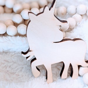 May include: A wooden unicorn cutout in a light brown color, sitting on a white fluffy surface. The unicorn is facing the right side of the image.