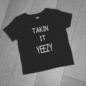 Peut inclure: T-shirt noir avec un texte blanc qui dit "Takin It Yeezy".