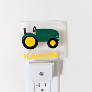 May include: A clear acrylic night light featuring a green tractor with yellow wheels. The name "KARSON" is printed in yellow below the tractor. The night light is plugged into a white wall socket.
