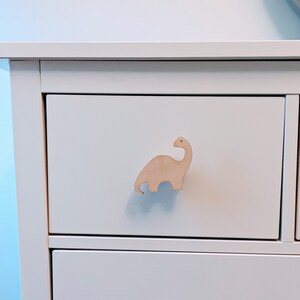 Peut inclure: Commode blanche à deux tiroirs. Le tiroir supérieur a un bouton en forme de dinosaure en bois.
