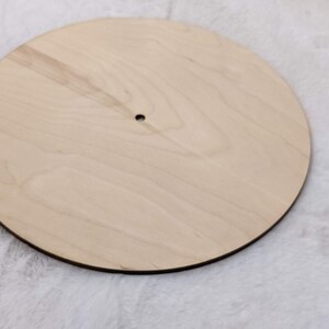 May include: A round, unfinished wooden disc with a small hole in the center. The wood has a light, natural color and a visible grain pattern. The edge is dark, creating a contrast with the light wood.