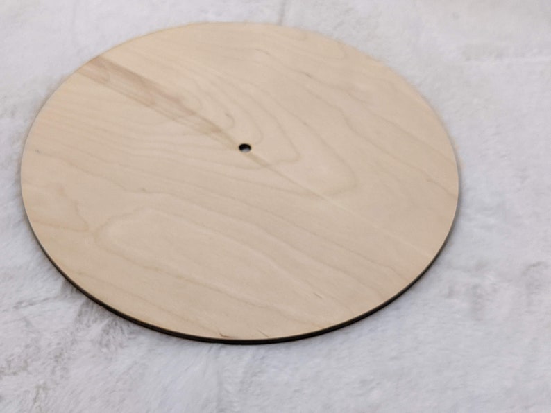 May include: A round, unfinished wooden disc with a small hole in the center. The wood has a light, natural color and a visible grain pattern. The edge is dark, creating a contrast with the light wood.