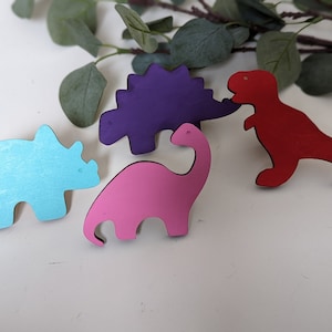 May include: Four wooden dinosaur shapes painted in different colors: blue, pink, purple, and red. The dinosaurs are arranged on a white surface.