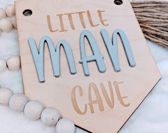 May include: Wooden sign with the text "Little Man Cave" in a light brown color with the word "Man" in a light blue color.
