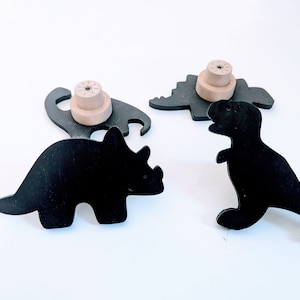 May include: Three black wooden dinosaur silhouettes with wooden knobs on the back. The dinosaurs are a triceratops, a stegosaurus, and a tyrannosaurus rex.