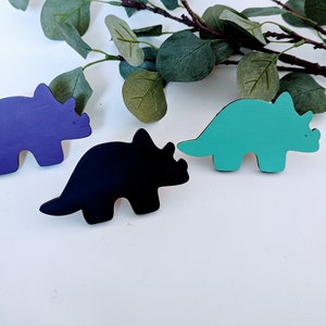 Peut inclure: Trois figurines de dinosaures en bois peintes en violet, turquoise et noir. Les dinosaures sont tous tournés dans la même direction et ont trois cornes sur la tête.