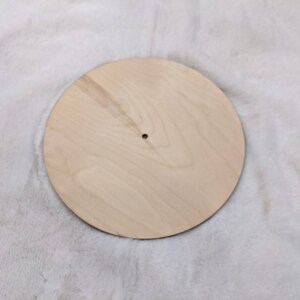 May include: A round, unfinished wooden disc with a small hole in the center. The disc is light-colored and appears to be made of plywood or a similar material. The wood grain is visible. The disc is likely a craft supply or a component for a larger project.