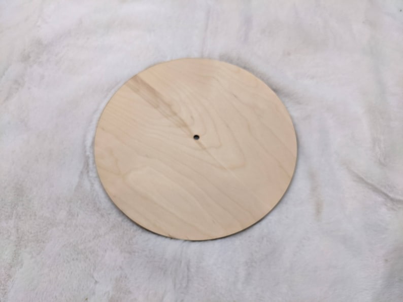 May include: A round, unfinished wooden disc with a small hole in the center. The disc is light-colored and appears to be made of plywood or a similar material. The wood grain is visible. The disc is likely a craft supply or a component for a larger project.