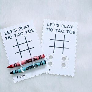 May include: Two white cards with scalloped edges, each with the text "Let's Play Tic Tac Toe" and a tic-tac-toe grid. One card has two circles with a number "1" below them. Two crayons, one red and one blue, are placed on the cards.