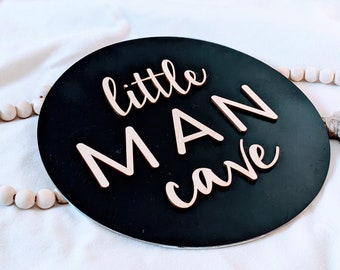 May include: Black round wooden sign with the text "little MAN cave" in white lettering. The sign is hanging from a white beaded garland with a brown tassel.