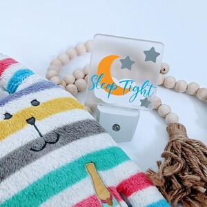 May include: A white night light with a clear acrylic front panel featuring a yellow crescent moon, three grey stars, and the text "Sleep Tight" in blue. The night light is next to a striped plush toy and a wooden beaded garland with brown tassels.