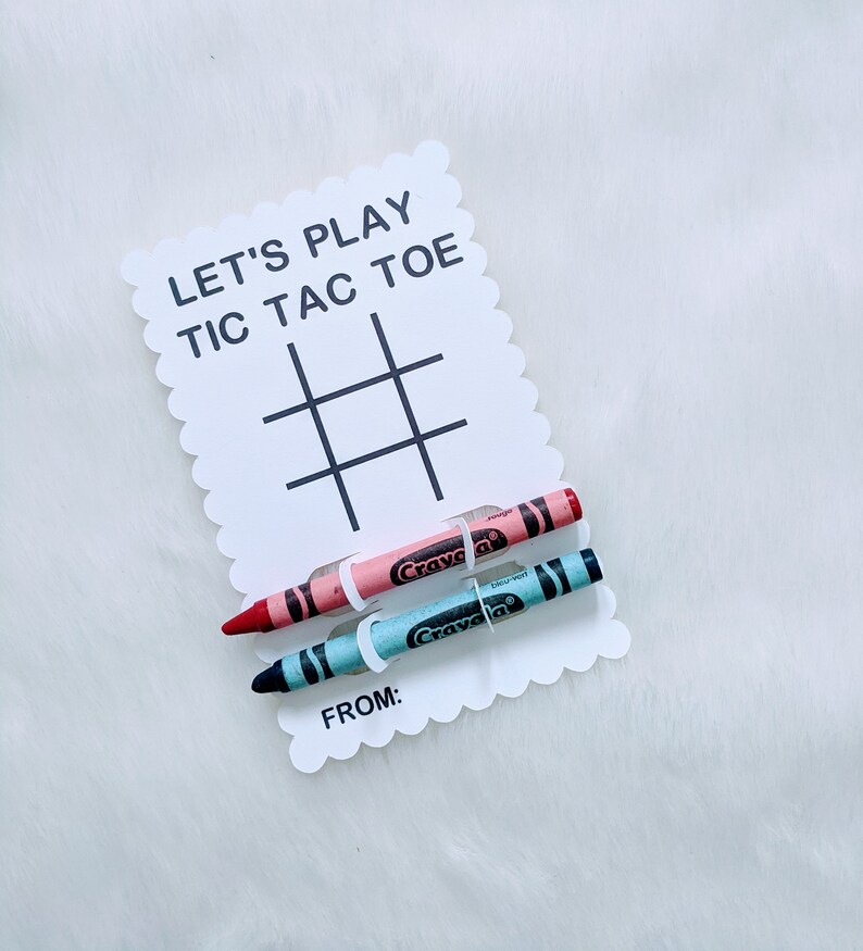 May include: A white card with the text "Let's Play Tic Tac Toe" and a tic-tac-toe grid. Two crayons, one pink and one blue, are attached to the card with white string.