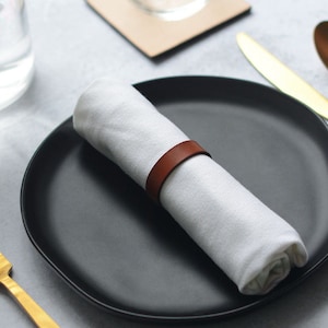 May include: A white napkin with a brown leather napkin ring is placed on a black plate. A gold fork and a gold knife are on the table.