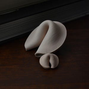 May include: Two off-white, folded fortune cookie-shaped objects made of a smooth, matte material. The larger object is positioned behind a smaller, identical version, both featuring a distinct curved, crescent-like shape with a central fold.