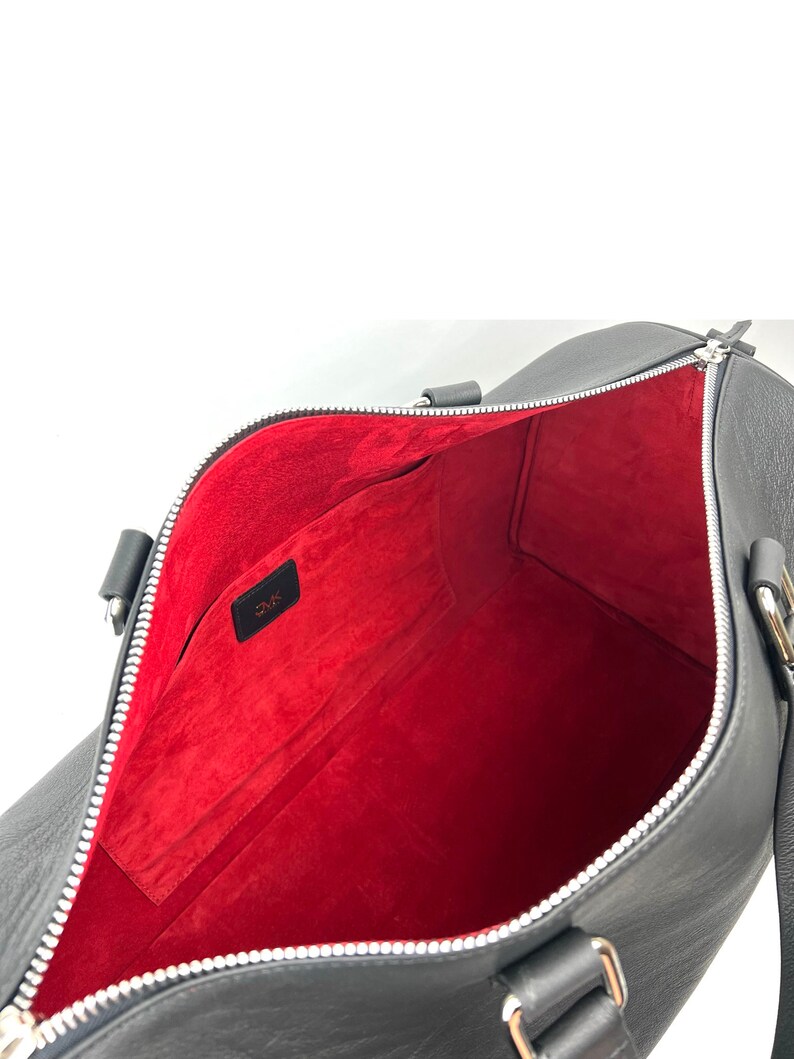 Puede incluir: Bolsa de viaje de cuero negro con forro interior rojo y cremallera plateada. La bolsa tiene un peque&ntilde;o bolsillo en el interior con la palabra "O.M.K." impresa en &eacute;l.