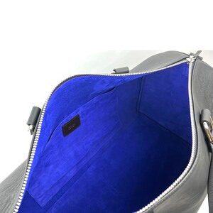 Puede incluir: Bolsa de viaje de cuero negro con forro de ante azul y cierre de cremallera. La bolsa tiene dos asas y un bolsillo con cremallera en el interior.