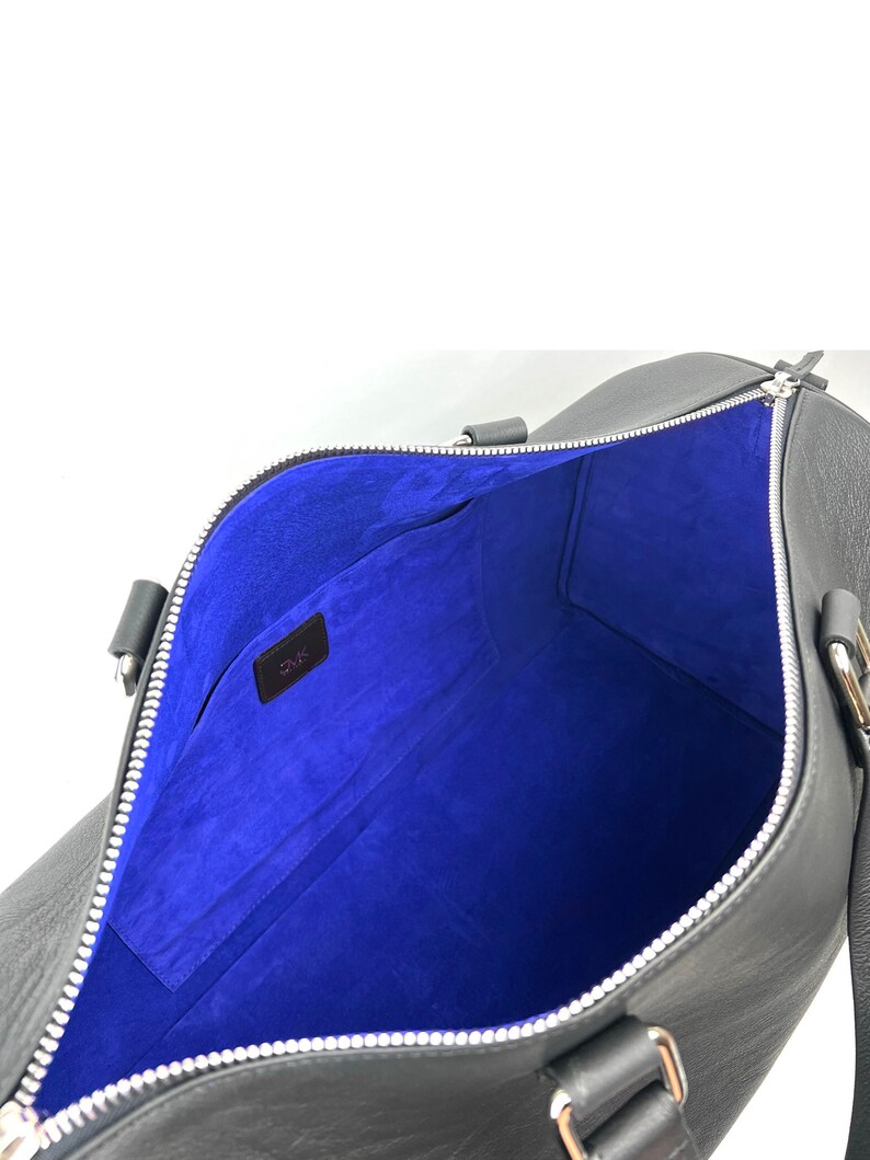 Puede incluir: Bolsa de viaje de cuero negro con forro de ante azul y cierre de cremallera. La bolsa tiene dos asas y un bolsillo con cremallera en el interior.