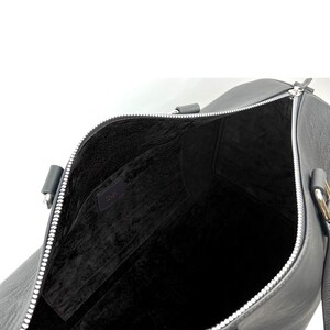 Puede incluir: Bolsa de viaje de cuero negro con cremallera plateada y forro de tela negro. La bolsa tiene dos asas y una correa para el hombro.