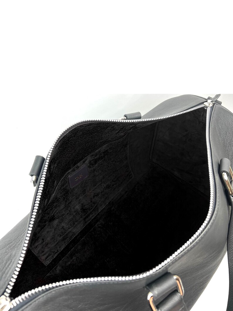 Puede incluir: Bolsa de viaje de cuero negro con cremallera plateada y forro de tela negro. La bolsa tiene dos asas y una correa para el hombro.