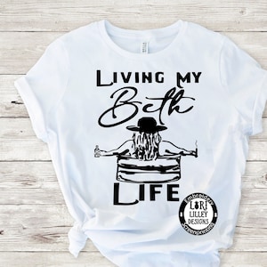 May include: White t-shirt with black text that reads "Living my Bath Life" and a black silhouette of a woman in a bathtub with a drink and a cigarette. The text "Embroidery Lori Lilley Designs Screenprinting" is in a circle below the image.