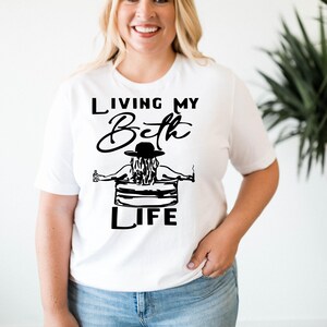 May include: A white short-sleeved t-shirt featuring a black graphic print. The design shows a person wearing a hat, sitting in a chair, holding a bottle and a cigarette, with the text "LIVING MY Beth LIFE" printed in black lettering above and below.