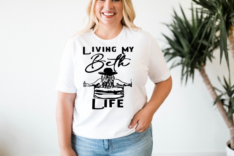 May include: A white short-sleeved t-shirt featuring a black graphic print. The design shows a person wearing a hat, sitting in a chair, holding a bottle and a cigarette, with the text "LIVING MY Beth LIFE" printed in black lettering above and below.