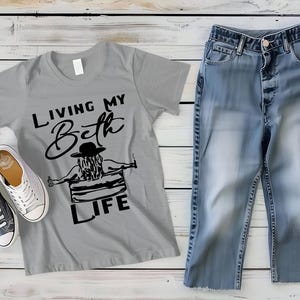 May include: A grey short-sleeve t-shirt featuring a black graphic of a person in a hat sitting in a tub with the text "LIVING MY Beth LIFE", paired with blue denim jeans and a pair of low-top black and white canvas sneakers with white laces.