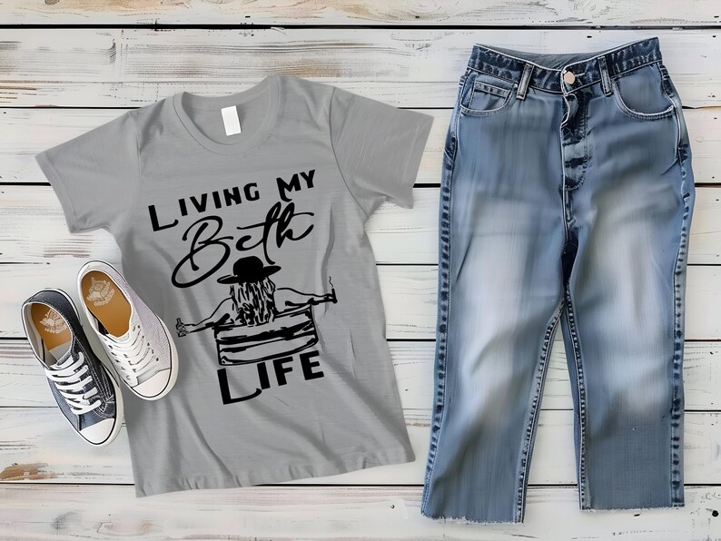 May include: A grey short-sleeve t-shirt featuring a black graphic of a person in a hat sitting in a tub with the text "LIVING MY Beth LIFE", paired with blue denim jeans and a pair of low-top black and white canvas sneakers with white laces.
