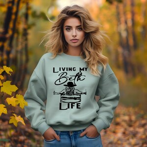 May include: A light sage green crewneck sweatshirt featuring a black graphic print of a person in a hat sitting in a tub with the text "LIVING MY Beth LIFE" printed in a combination of bold sans-serif and cursive black lettering on the front.