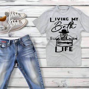 May include: A light gray short-sleeved t-shirt with a black graphic of a person in a hat sitting in a tub, featuring the text "LIVING MY Beth LIFE," paired with blue denim jeans and a pair of light gray lace-up canvas sneakers with white soles.