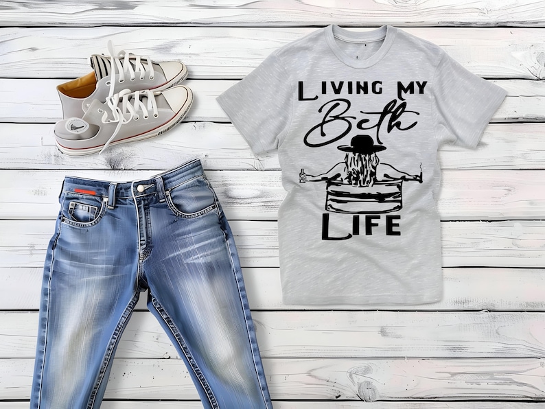 May include: A light gray short-sleeved t-shirt with a black graphic of a person in a hat sitting in a tub, featuring the text "LIVING MY Beth LIFE," paired with blue denim jeans and a pair of light gray lace-up canvas sneakers with white soles.
