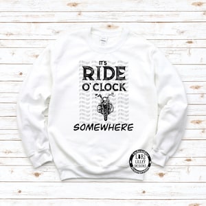 May include: White sweatshirt with black text that reads "It's Ride O'Clock Somewhere" with a motorcycle graphic. The text is repeated in a smaller font in the background.