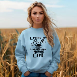 May include: A light blue crewneck sweatshirt featuring a black graphic print on the chest. The design depicts a person wearing a hat, sitting in a tub, with the text "LIVING MY Beth LIFE" printed in a combination of block and script lettering.