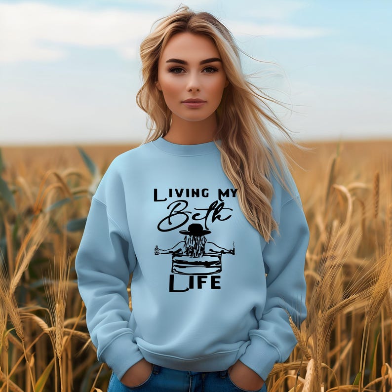 May include: A light blue crewneck sweatshirt featuring a black graphic print on the chest. The design depicts a person wearing a hat, sitting in a tub, with the text "LIVING MY Beth LIFE" printed in a combination of block and script lettering.