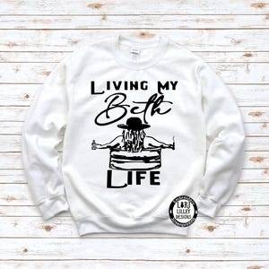 May include: A white long-sleeve sweatshirt featuring a black graphic print. The design shows a person in a hat sitting in a tub, holding a bottle and a cigarette, with the text "LIVING MY Beth LIFE" and a small circular logo in the corner.