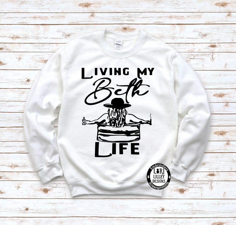 May include: A white long-sleeve sweatshirt featuring a black graphic print. The design shows a person in a hat sitting in a tub, holding a bottle and a cigarette, with the text "LIVING MY Beth LIFE" and a small circular logo in the corner.