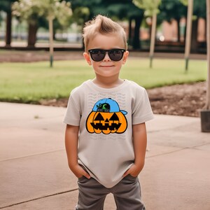 May include: A young child wearing a light gray t-shirt with a pumpkin wearing a blue baseball cap graphic. The pumpkin has a black and green face with a toothy grin. The child is wearing black sunglasses and gray pants.