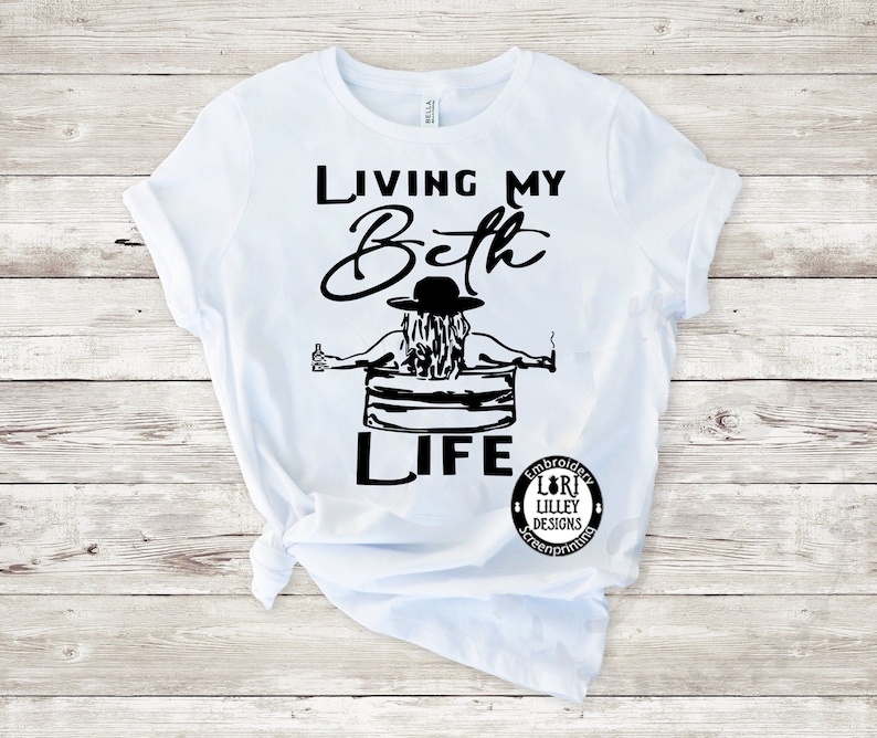 May include: A white short-sleeve t-shirt featuring a black graphic of a person in a hat sitting in a tub with a drink and a cigarette. The text reads "LIVING MY Beth LIFE" with a circular logo that says "EMBROIDERY LORI LILLEY DESIGNS SCREENPRINTING".