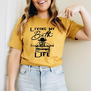May include: A mustard yellow short-sleeve t-shirt featuring a black graphic print of a person wearing a hat while sitting in a tub. The text reads "LIVING MY Beth LIFE" in a combination of bold block lettering and a cursive script font.