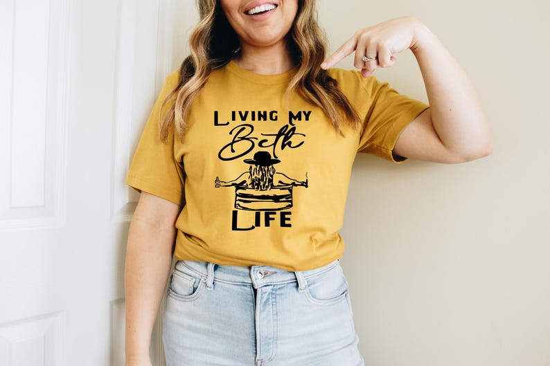 May include: A mustard yellow short-sleeve t-shirt featuring a black graphic print of a person wearing a hat while sitting in a tub. The text reads "LIVING MY Beth LIFE" in a combination of bold block lettering and a cursive script font.