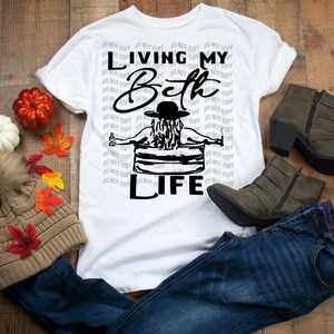 May include: A white short-sleeved t-shirt featuring a black graphic of a person in a hat sitting in a tub with the text "LIVING MY Beth LIFE". The shirt is displayed with a tan knit beanie, blue jeans, and dark brown ankle boots.